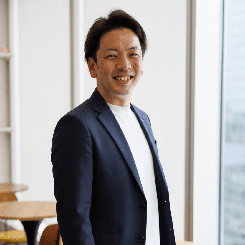 編集済 株式会社デジタルブロックス 池部 拓也 | テクロ株式会社 Smiling man in a navy blazer and white shirt standing by a large window in a bright office setting.