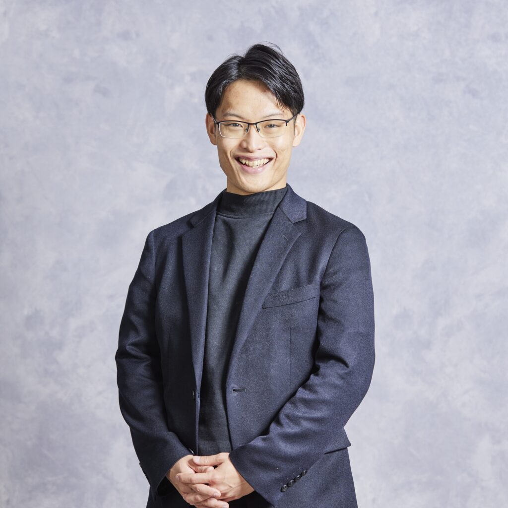 編集済 代表取締役 天野 央登 | テクロ株式会社 Smiling man in a dark blazer and black turtleneck with hands clasped in front, against a gray textured background.